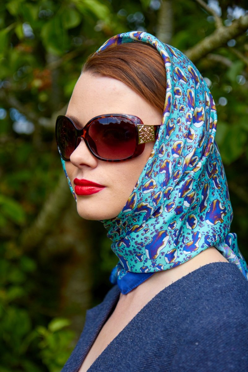 Silk Head Scarf - Elegant woman wearing a silk head scarf adorned with a delicate raindrops pattern. The scarf, featuring various shades of blue, wraps gracefully around her head and neck. She has large, dark sunglasses and a serene expression, complemented by her bold red lipstick. The background is a soft blur of greenery, enhancing the sophisticated and natural aesthetic of the image.
